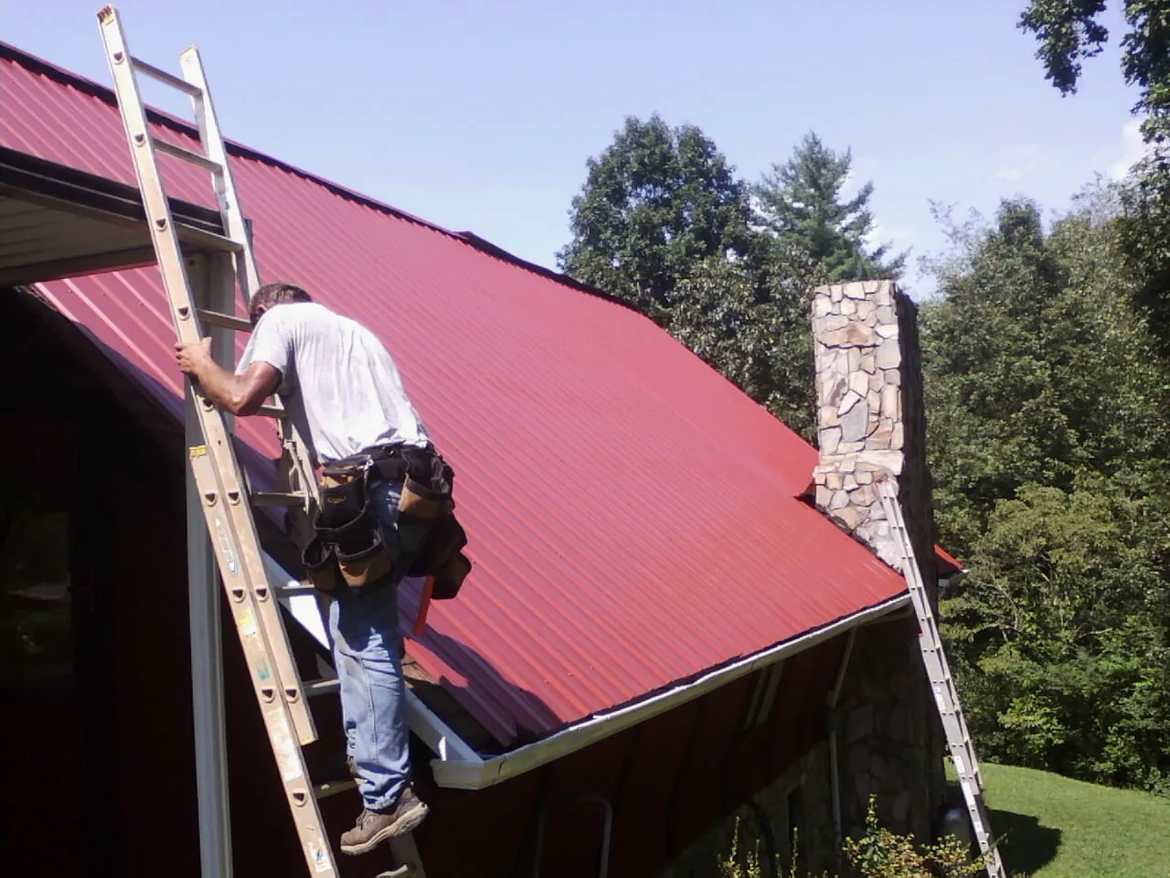 Roofer on a ladder beside a red metal roof and stone chimney — Soffit & Fascia Repair by Roofing Brothers in Prospect Hill